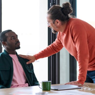 Senior boss motivating employee in front of his colleagues at meeting in office. Executive saying good word to his employee sitting at desk touching him with hand.