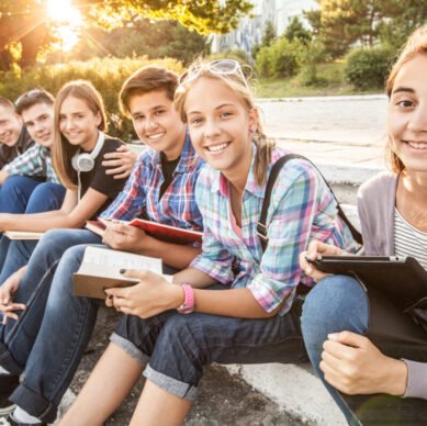 group of young students with books and gadgets sit on the steps in the park