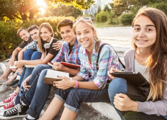 group of young students with books and gadgets sit on the steps in the park